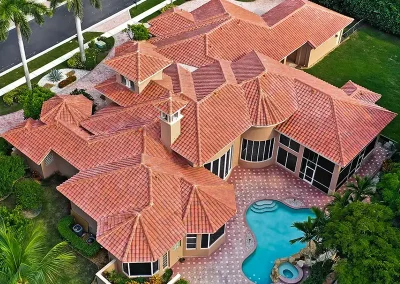 inf-01 - Infinity Roofing - A Stronghouse Partner Aerial view of a luxury villa with multiple red-tiled roofs and a curved blue pool surrounded by palm trees.