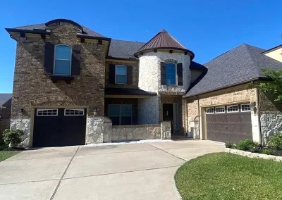 inf-04 - Infinity Roofing - A Stronghouse Partner Two-story suburban home with a stone turret, brick facade, and two attached garages under a clear blue sky.