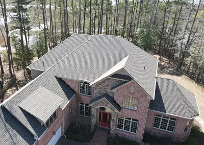 inf-05 - Infinity Roofing - A Stronghouse Partner Aerial view of a large brick house with a red front door, stone entry surround, multiple gabled rooflines, and a two-car garage, surrounded by trees