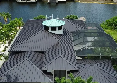 inf-07 - Infinity Roofing - A Stronghouse Partner Aerial view of a large house with dark metal roofs and a turquoise cupola, with a screened pool enclosure to the right and a canal behind.